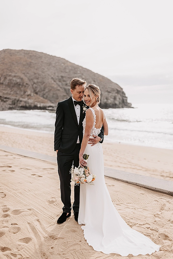 Couple portrait at a beach wedding portrait, bride in low-back lace-strap gown with bouquet embracing groom in black tux by ocean cliffs