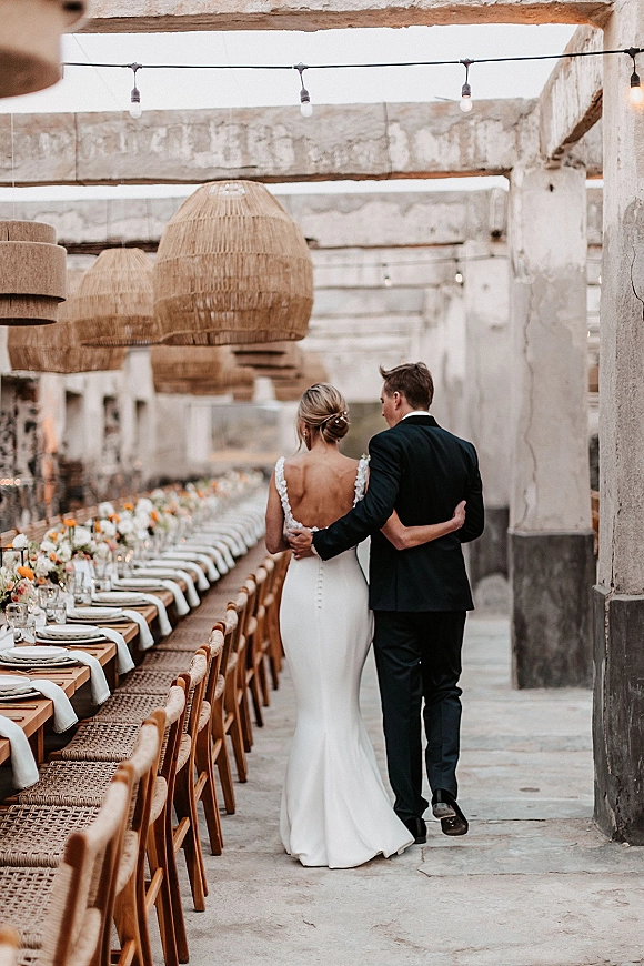 Couple portrait of bride and groom walking away, arm around waist, past a long banquet table under string lights at an industrial venue