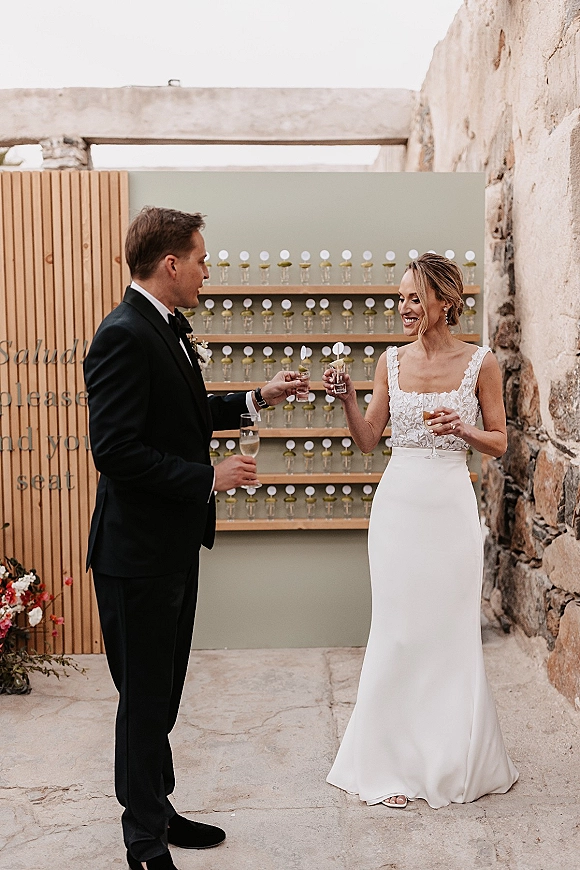 Wedding toast as bride and groom clink glasses in lace dress and black tux, with escort card display and stone courtyard wall behind