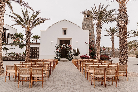 Ceremony setup with outdoor wedding ceremony seating of wood cane-back chairs facing a floral arch at a white chapel in a desert courtyard