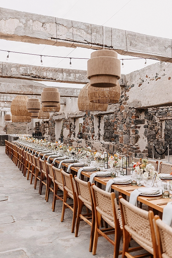 Reception tablescape with long wedding table set with white runner, pastel florals, candles and black lanterns under wicker pendants in stone ruins courtyard