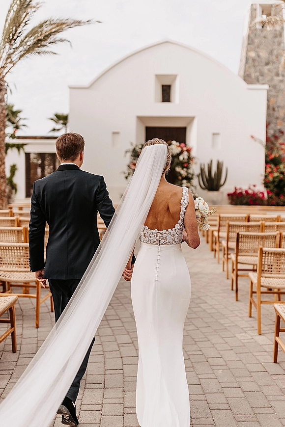 Wedding processional with bride walking down aisle in a long veil holding a bouquet beside groom, toward a floral arch in a courtyard chapel