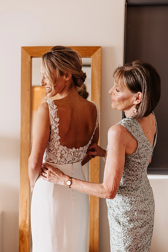 Bride getting ready as her mother fastens wedding dress buttoning on a low back lace gown, seen in a wooden mirror by window light
