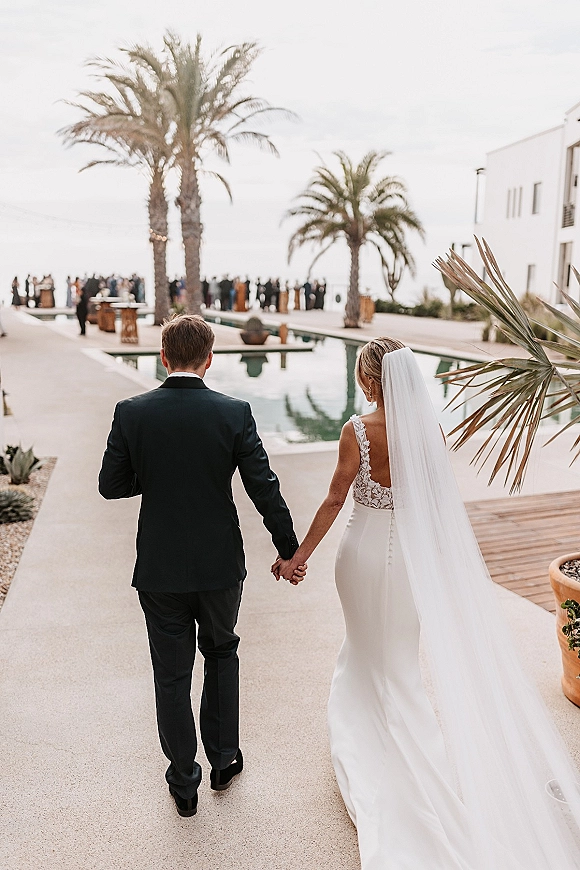 Processional moment as bride and groom walking hand in hand, veil trailing, past poolside guests with palms, ocean and modern venue behind