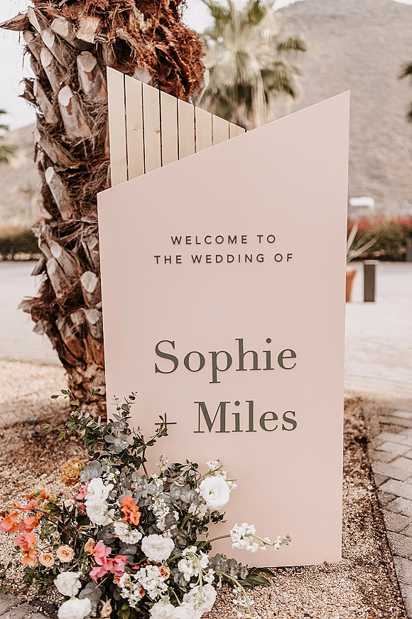 Wedding welcome sign with serif typography on a freestanding board, accented by eucalyptus and peach-white flowers, set by desert plants and mountains