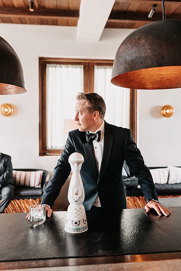 Groom portrait in a black tuxedo, leaning on a bar counter with a rocks glass and ceramic decanter in a lounge with pendant lights