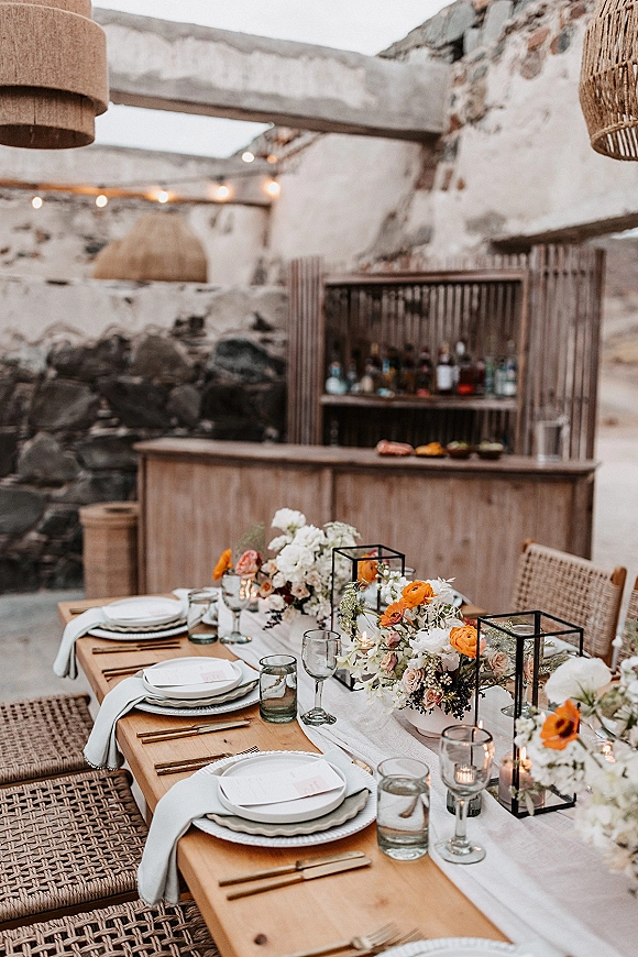 Reception tablescape with wedding table runner on a wooden banquet table, orange and white florals, black lanterns, candles, and string lights near a stone wall bar