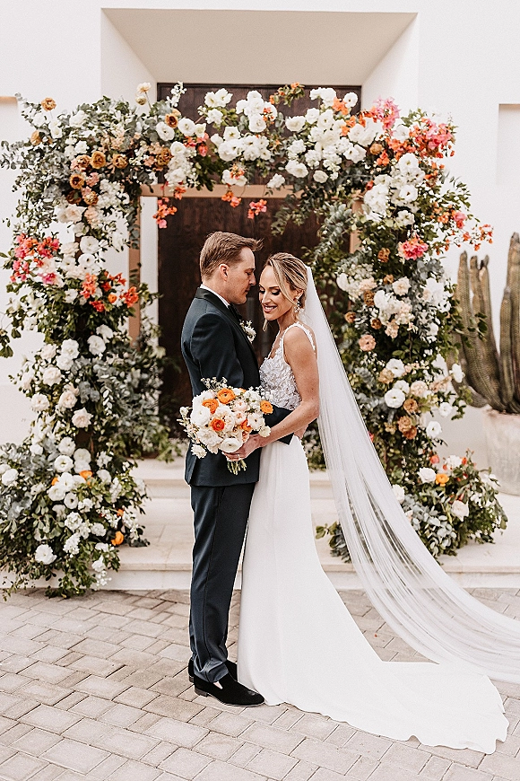Couple portrait of bride and groom embrace, her lace dress and long veil by a floral arch on stone steps in a desert courtyard