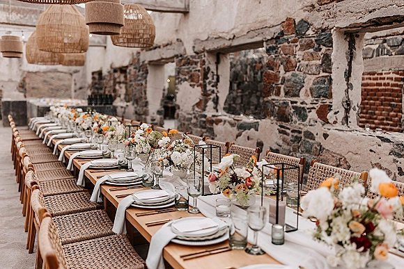 Reception tablescape with a long banquet table, white runner, floral bud vases, black lanterns, and candlelight under wicker pendant and string lights.
