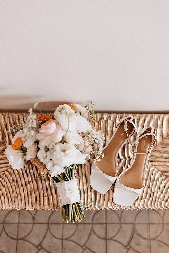 Bridal details with wedding detail flat lay of white strappy heels and a blush, orange, and white bouquet on a woven bench by a white wall