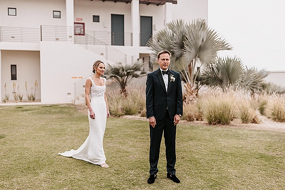 First look moment as the bride in a fitted dress approaches a groom in black tuxedo on a palm-lined lawn by a modern staircase