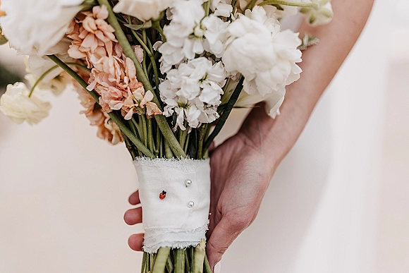 Bridal bouquet of white and peach flowers with pearl-pinned wrap and a ladybug, held against a white dress with blurred greenery behind