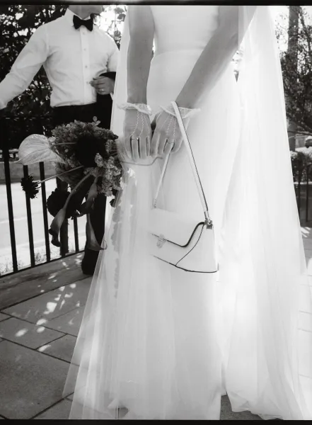 Bride portrait in a black and white wedding portrait, holding bouquet and handbag in lace gloves beside groom on a sunlit patio railing