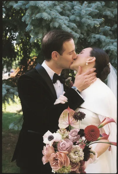 Wedding kiss portrait of bride and groom kissing, groom’s hand on her face as she holds a bouquet in an evergreen garden backdrop
