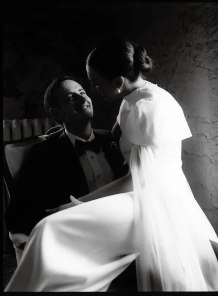 Couple portrait in black and white, bride sitting on groom’s lap on an upholstered bench, leaning in as he looks up by a textured wall