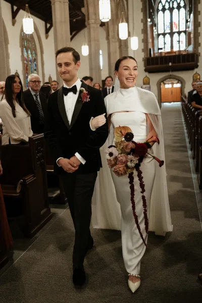 Wedding recessional as bride and groom walk the church aisle, she holds a bouquet and wears a cape dress and veil amid pews and stained glass