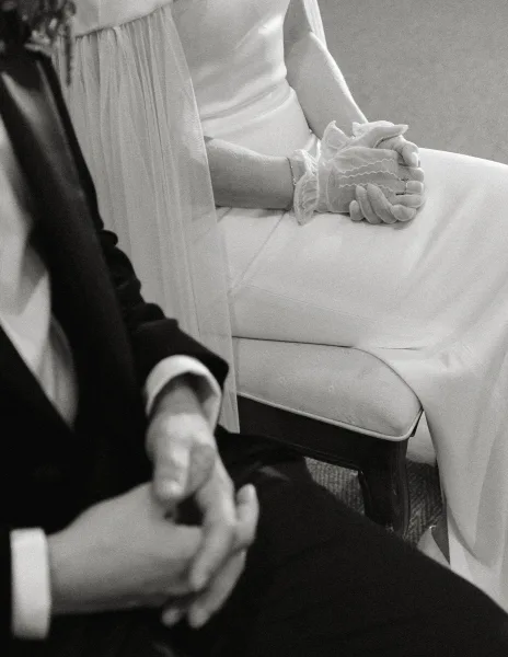 Ceremony moment as bride and groom sit with clasped hands, lace-gloved bride showing wedding rings on an indoor carpeted aisle