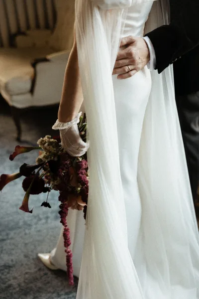 Couple portrait of bride and groom embrace, his hand on her waist as her long veil drapes and calla lily bouquet trails in an indoor room