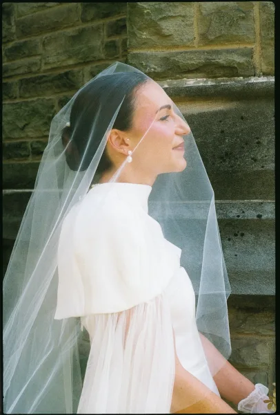 Bridal portrait of a bride in side profile wearing a wedding veil and pearl drop earrings, hair in a low bun beside a stone wall