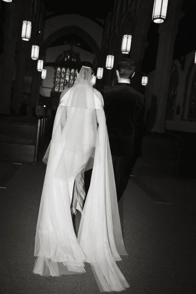 Ceremony processional as bride walking down aisle beside groom, long veil trailing over runner in church with stained glass and lanterns