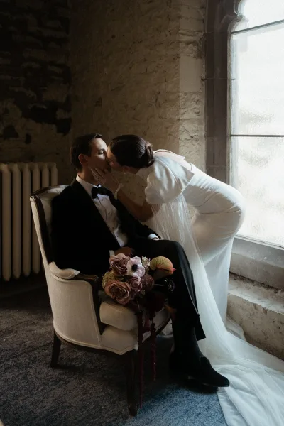 Wedding kiss portrait of bride leaning in to kiss seated groom in black tuxedo, her veil over shoulder, bouquet on lap by stone wall window