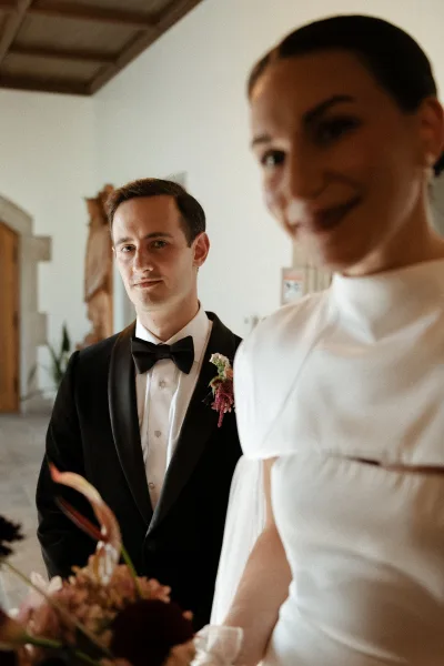 Couple portrait of bride in high-neck dress holding a burgundy bouquet beside groom in black tuxedo, with chapel altar cross behind them