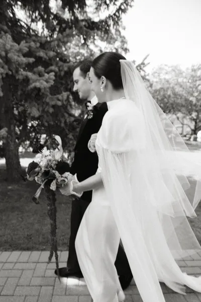 Wedding couple portrait in a black and white wedding photo, bride holding bouquet with veil trailing as they walk along a tree-lined path