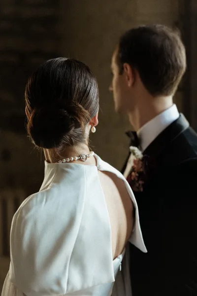 Couple portrait from behind showing bride’s sleek low bun and pearl earrings beside groom in black tuxedo against a dark wall