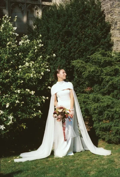 Bridal portrait of a bride in a wedding dress with cape, holding a bouquet with anthurium, standing by garden shrubs and white roses