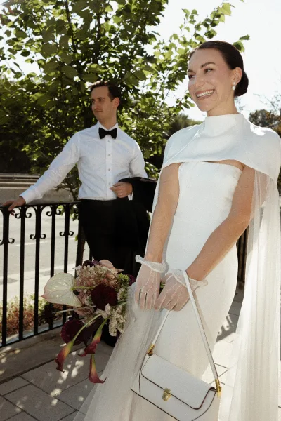 Bridal portrait of a bride in a minimalist wedding dress with cape overlay, holding a bouquet on a sunlit terrace with iron railing and trees