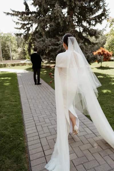 First look moment as bride walking to groom with long veil train and calla lily bouquet behind her back on garden walkway