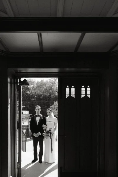 Wedding couple portrait of bride in veil holding a bouquet with ribbon accents beside groom in tuxedo at a doorway with outdoor steps