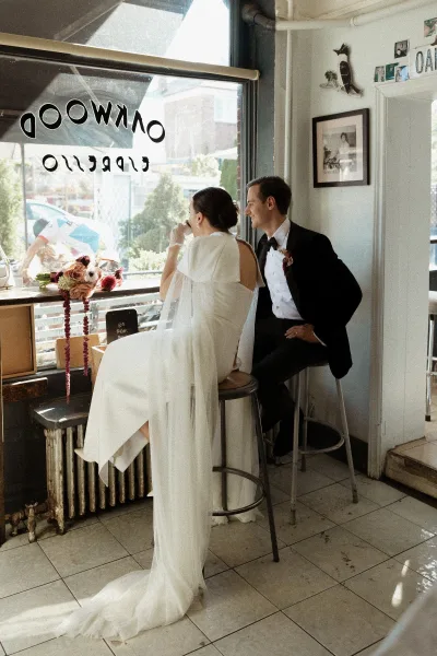 Couple portrait of bride and groom at cafe, seated on bar stools by a large window; bride holds a burgundy bouquet as groom watches