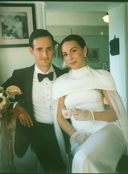 Couple portrait of bride in cape veil holding a coffee cup beside groom in tuxedo, standing in a doorway with framed photos behind