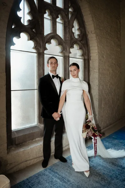 Couple portrait of bride and groom holding hands, bride in cape gown with gloves and bouquet, by gothic arched windows indoors
