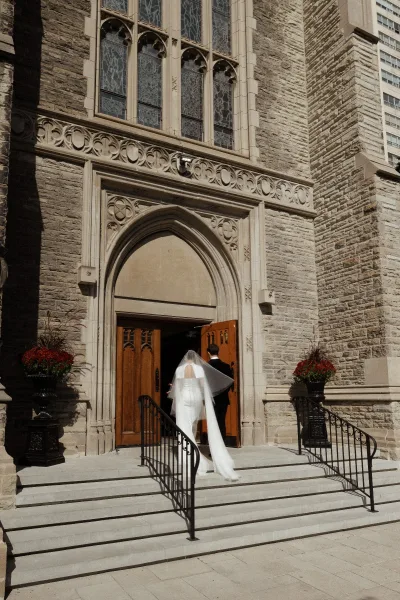 Ceremony entrance as bride walking into church with long veil and gown train beside groom in black tuxedo at stone arched doorway
