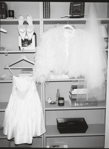 Wedding dress on a bridal dress hanger with veil accent, styled with shoes and pearl earrings beside a bookshelf in a home interior
