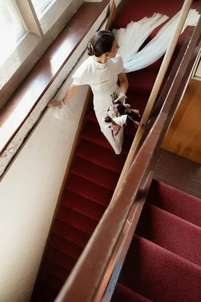 Bridal portrait of bride on staircase holding a burgundy and blush calla lily bouquet, veil train on red carpet stairs in window light