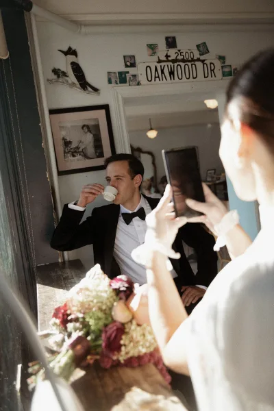 Groom portrait in a black tuxedo and bow tie sipping an espresso by window light, reflected in a mirror in a cozy prep room