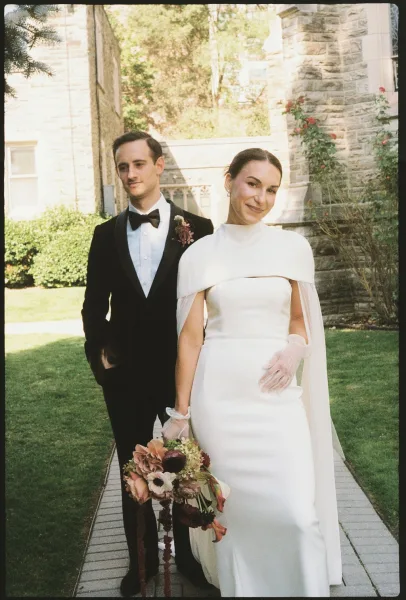 Wedding couple portrait of bride in a white cape-sleeve gown and sheer gloves holding bouquet beside groom in black tuxedo on stone walkway
