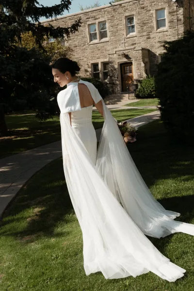 Bridal portrait of a bride in a modern bridal gown with a long train, holding a bouquet on a lawn by a stone building and trees