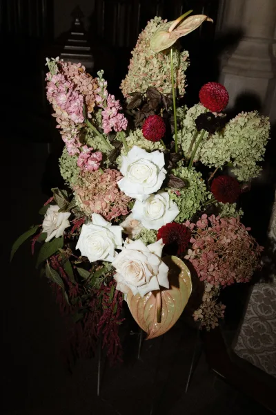 Wedding floral arrangement with white roses and hydrangea blooms, anthurium and red dahlias against a dark interior wall and stone column