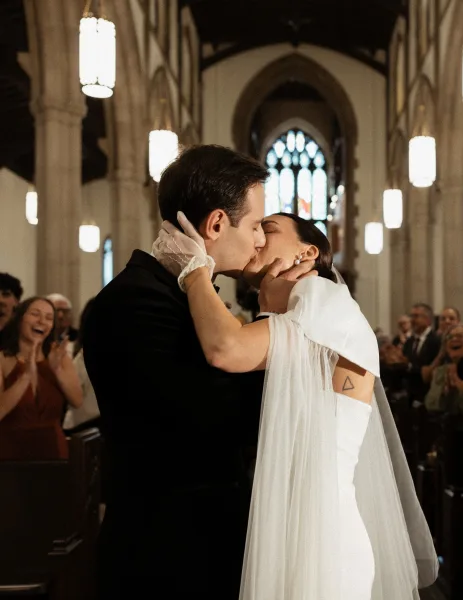 Wedding kiss as bride and groom share their first kiss in a church aisle, her veil flowing beneath stained glass as guests cheer
