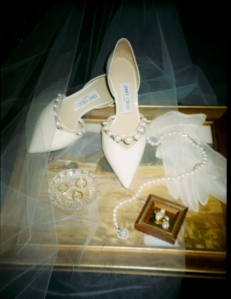 Bridal shoes and pearl wedding heels on a marble tray with tulle veil, ring set in crystal dish, and pearl earrings on dark fabric