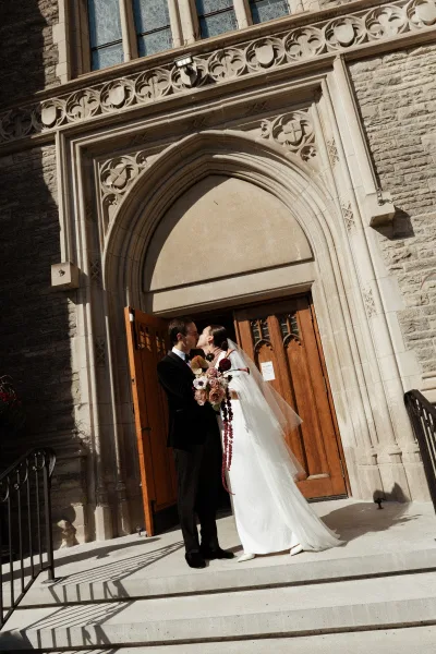 Wedding kiss as bride in a simple dress holds a bouquet, long veil flowing outside a stone church entrance with arched doors