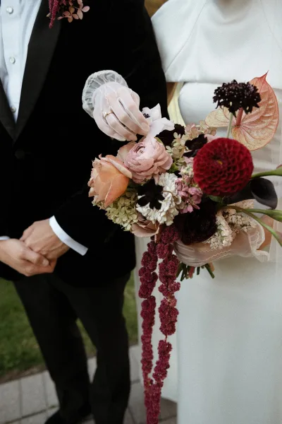 Bridal bouquet in burgundy wedding bouquet tones with roses, anthurium, dahlias and trailing amaranthus, held by gloved hands on an outdoor walkway