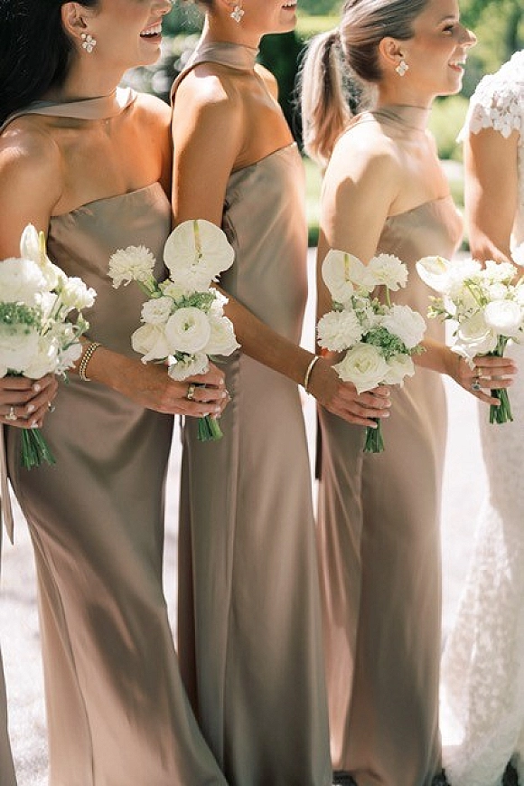 Bridesmaid dresses in taupe bridesmaid dresses, long satin halter gowns with white rose bouquets and greenery on a sunlit walkway
