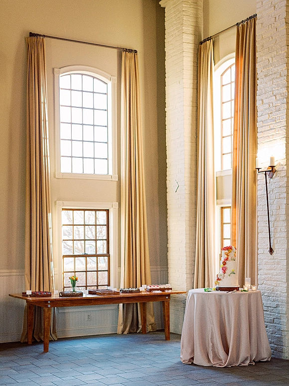 Wedding cake table with a tiered buttercream cake and sugar flowers, flanked by candles on a round table near tall windows and brick wall