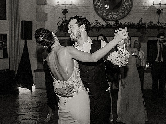 First dance as bride in a low-back wedding dress and groom in vest slow dance by candlelit fireplace, guests watching near brick wall