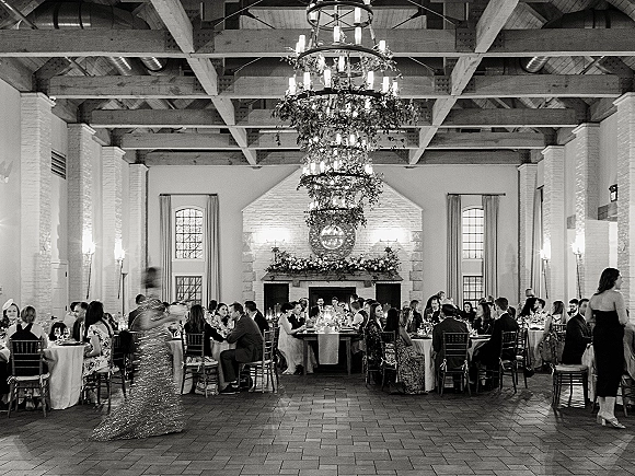 Wedding reception with candlelit tables and chandelier, long banquet and round settings around a white brick fireplace in a hall